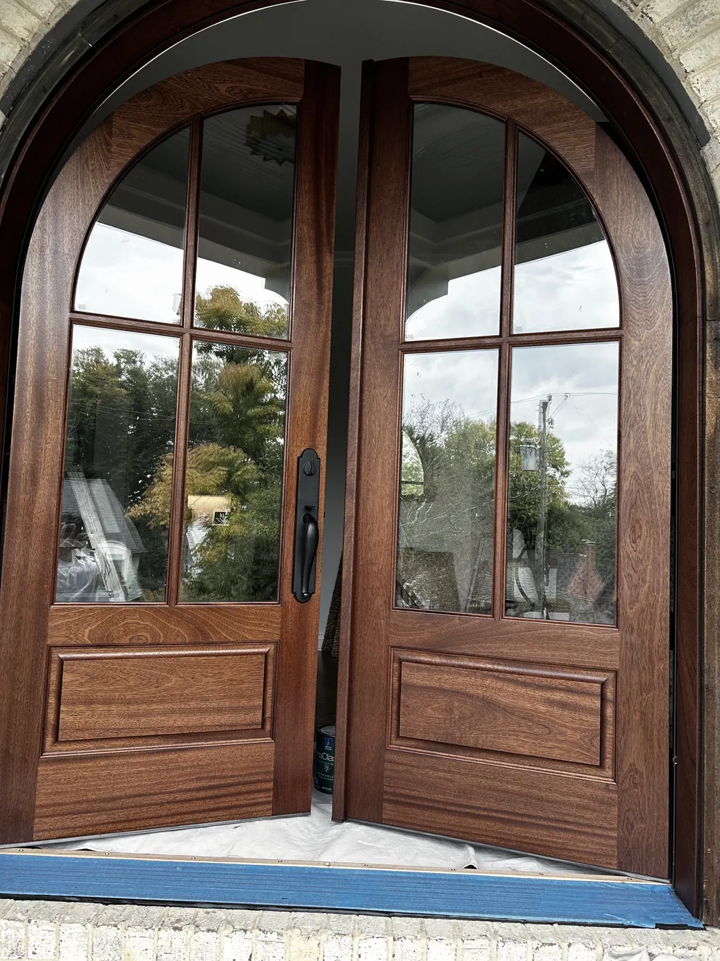 Arched pair of wood double doors with glass panes and black handles in a pale brick archway, the wood a deep red-brown, trees reflected in the glass.