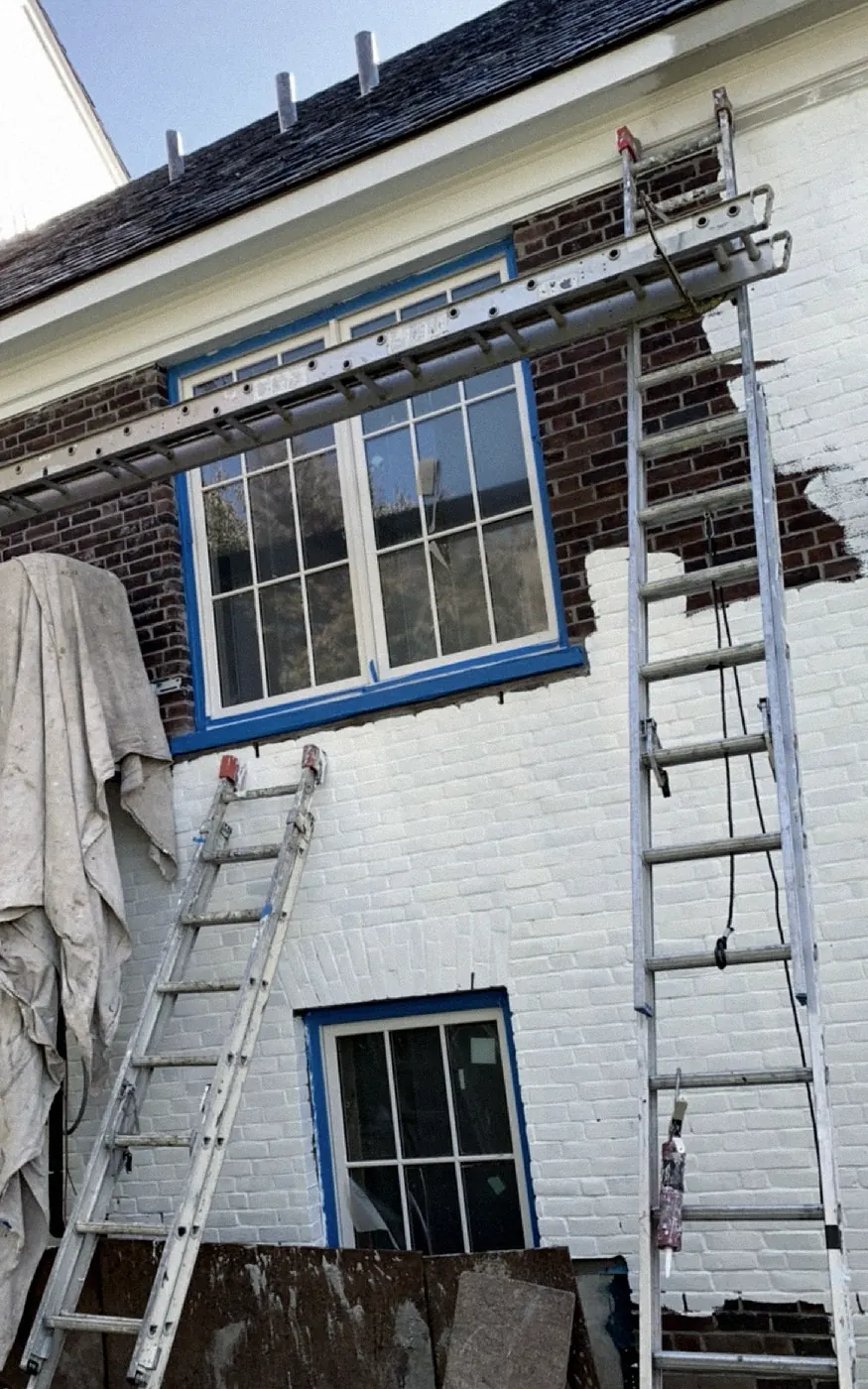 During: brick painted white from the bottom up, dark red-brown brick still showing around a blue-taped window, extension ladders and a hanging drop cloth.
