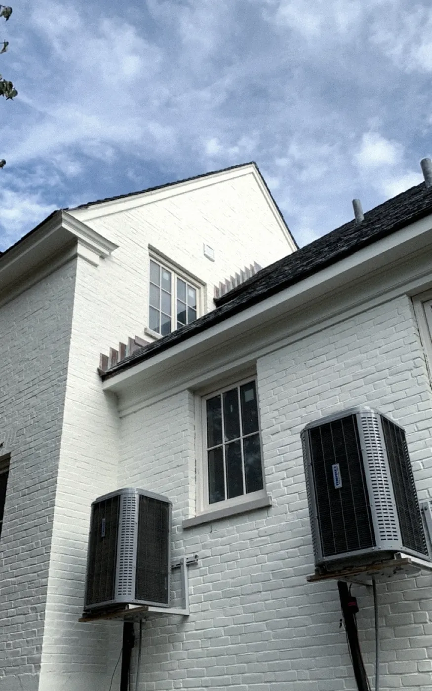 After: a brick wall painted white with cream window trim, two air conditioner condensers on brackets and a gabled roofline against a blue sky.
