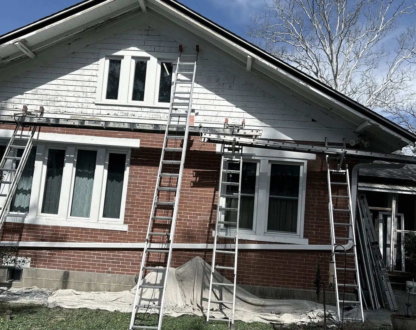 During: extension ladders against a brick bungalow, drop cloths along the foundation, the gable scraped.