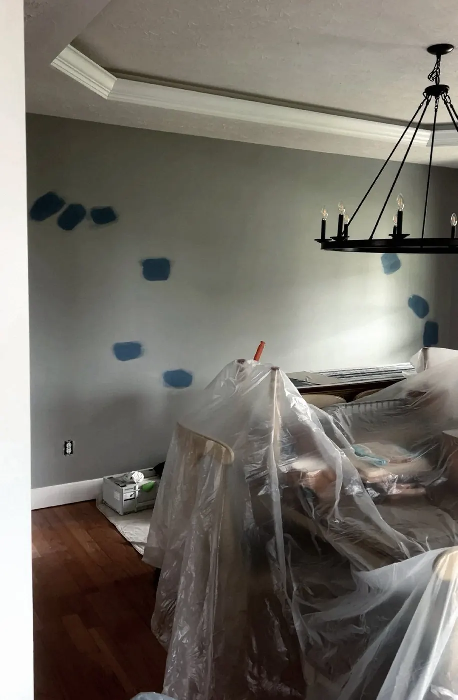 Dining room mid-job: furniture wrapped in plastic under an iron chandelier, blue paint swatches on the gray wall.