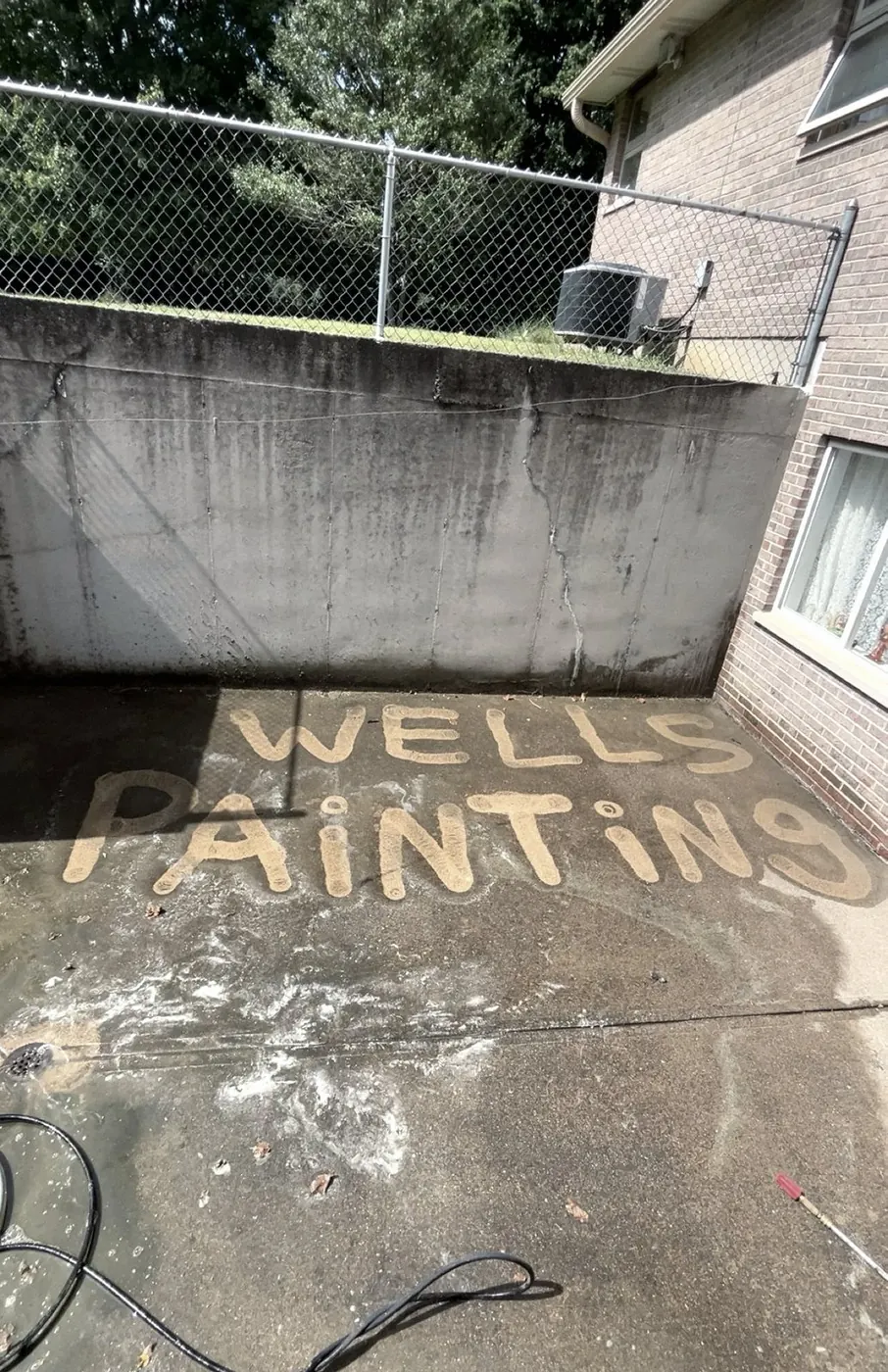 Concrete patio where the words WELLS PAINTING have been cleaned into the grime, a hose and a retaining wall behind.
