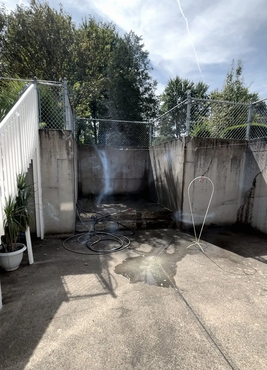 Concrete patio inside stained retaining walls and a chain-link fence, a spray wand and hose on the ground, a mist plume and a puddle.