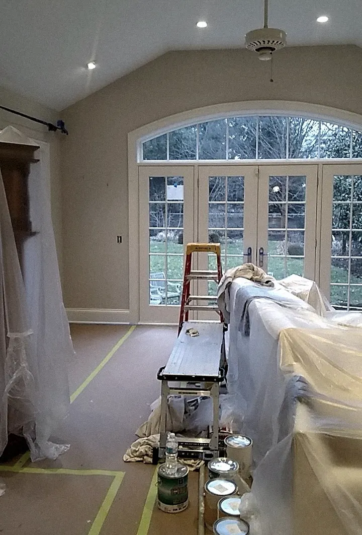 Vaulted room with an arched window over French doors, the floor covered in brown paper with green tape lines, furniture wrapped in plastic, a stepladder and a row of paint cans.